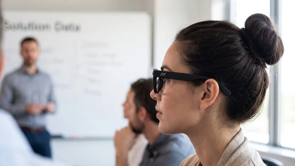 A woman wearing AURVIZ o1 smartglasses listens to a colleague presenting at a whiteboard in a meeting room.