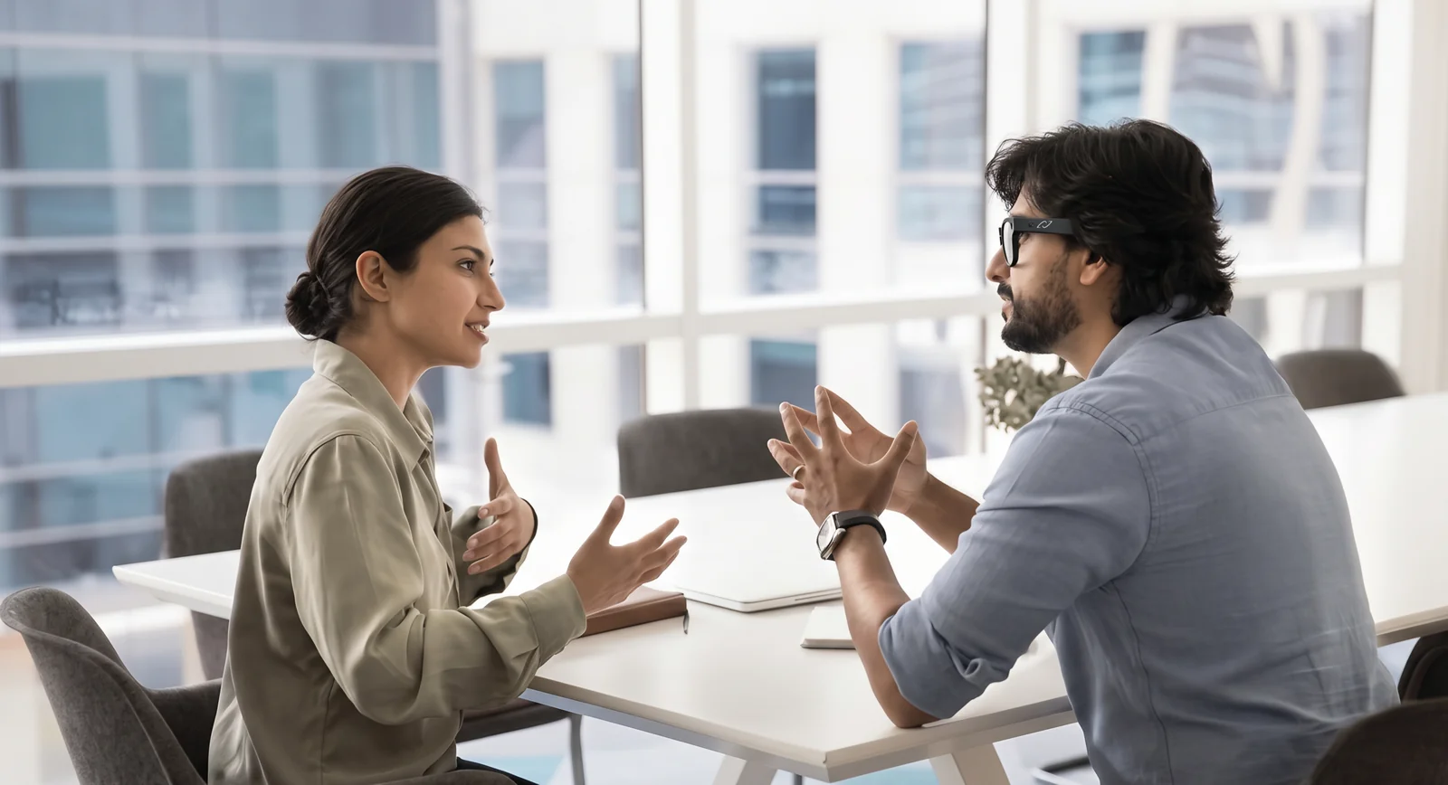 A salesperson wearing AURVIZ O1 glasses talks with a client across a table in a bright office