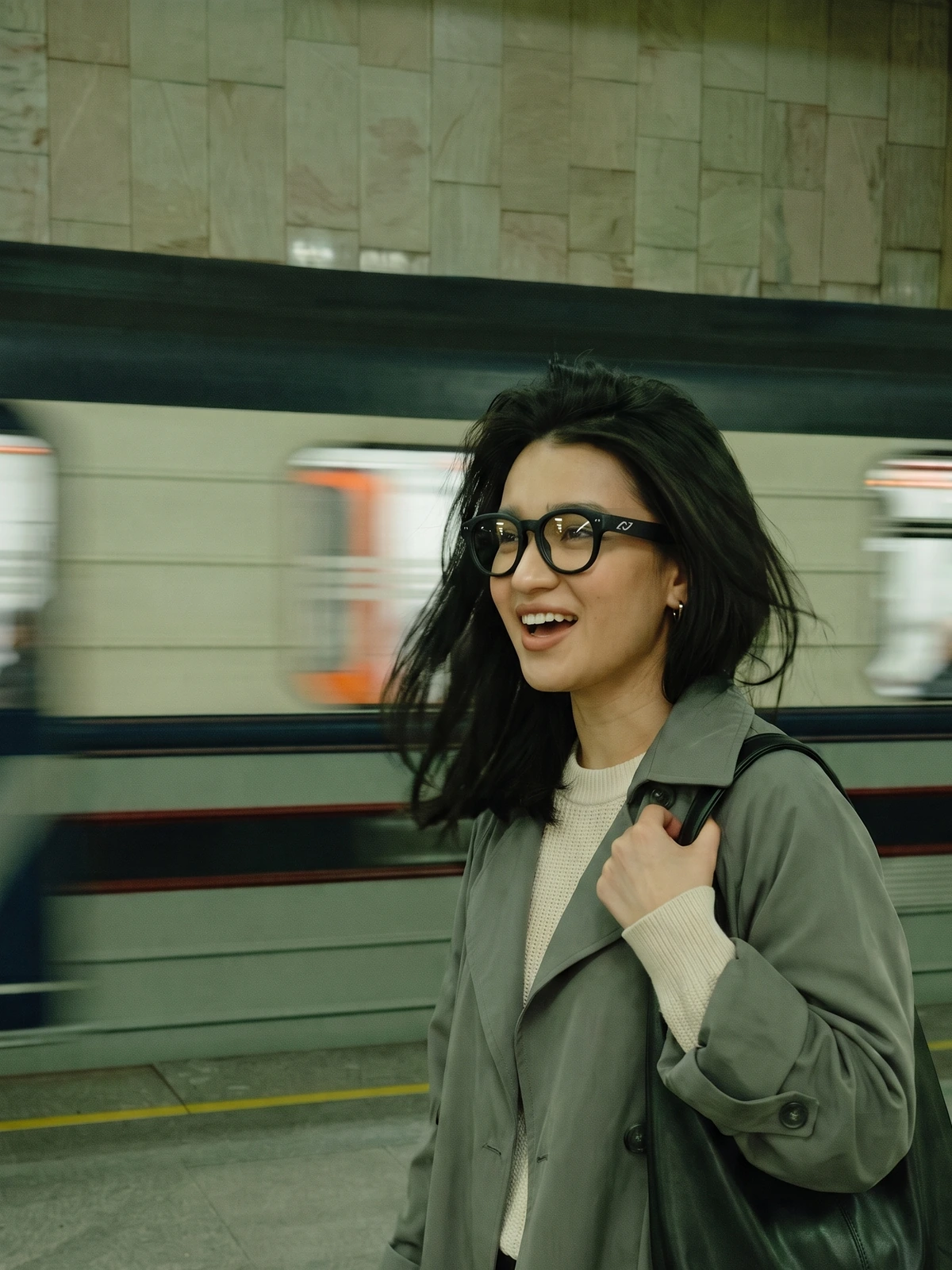 A woman on an underground platform, speaking mid-call, wearing the AURVIZ o1 smartglasses as a train rushes past behind her
